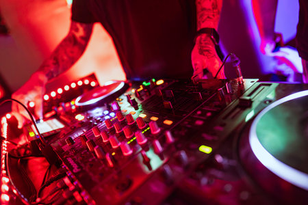 Rovigo, Italy 18 December 2025: dj hands with tattoos operating a mixing console and turntable with red and colorful lights in a lively nightclub settingのeditorial素材