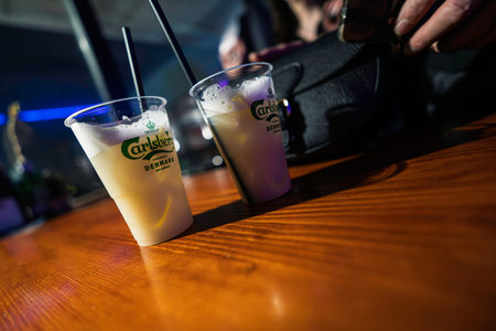 Rovigo, Italy 18 December 2025: two Carlsberg beer cocktails in plastic cups with straws on a wooden bar counter at a lively nighttime party, bokeh lights illuminating foamのeditorial素材