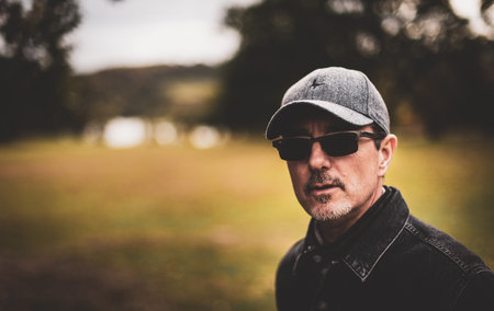 A white caucasian man in a baseball hat and sunglasses looks directly at the camera
