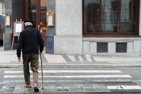 An elderly male with a walking stick slowly walks across a crosswalk in the middle of a city aloneの写真素材