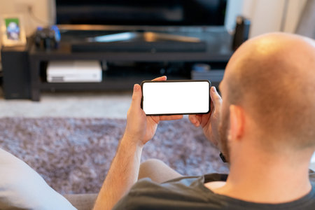 A white Caucasian male holds a black smartphone with a big white screen in a moden day living room...