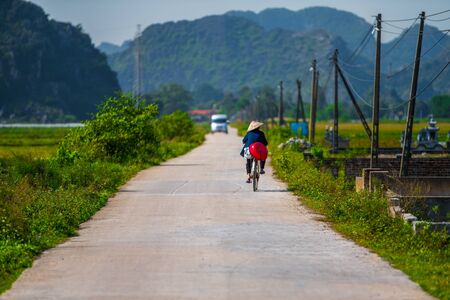 A local Tam Coc resident rides along a small road beside Rice Fields and beautiful mountain landscapes in Ninh Binh, Northern Vietnam - Autumn 2019の写真素材