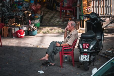 Hanoi, Vietnam - 12th October 2019: An old elderly Asian man sits on a plastic chair next to a moped on the phoneのeditorial素材