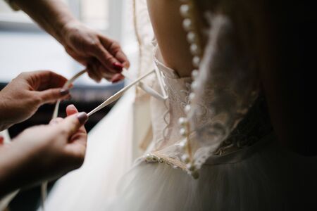 A white wedding dress is knitted to the bride. A bride is being helped to wear the wedding dress. The bride's preparation for the wedding