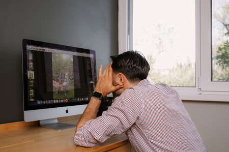 Tired photographer works in Photo Editing App. Software on His Personal Computer.Photographer for processing photos. Photographer equipment on the table. Retouching photos.のeditorial素材