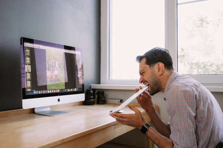 angry photographer works in Photo Editing App. Software on His Personal Computer.Photographer for processing photos. Photographer equipment on the table. Retouching photos.のeditorial素材