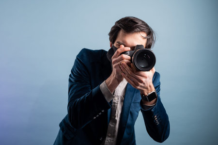 Portrait of positive photographer, man with medical mask holding professional digital dslr camera. Indoor studio shot isolated on blue background. The photographer is wearing a medical mask.の写真素材