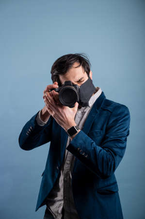 Portrait of positive photographer, man with medical mask holding professional digital dslr camera. Indoor studio shot isolated on blue background. The photographer is wearing a medical mask.の写真素材