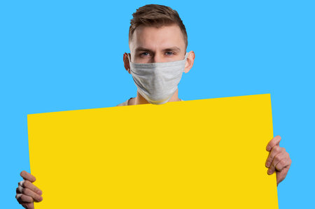 Young man wearing medical mask showing yellow paper poster and looking at camera on blue...