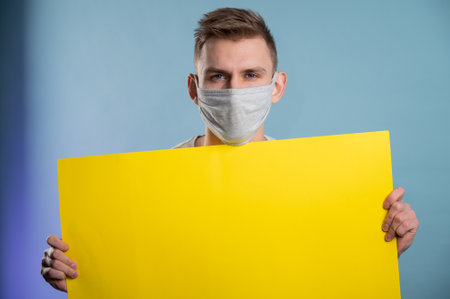Young man wearing medical mask showing yellow paper poster and looking at camera on blue background.Advertisement Conceptの写真素材