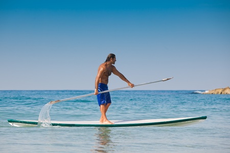 The guy with an oar on a surfboard. The beautiful brawny guy with an oar in hands on a surfboard.の写真素材