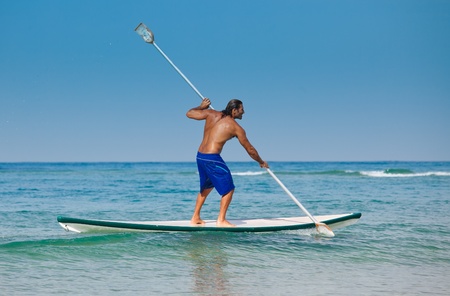 The guy with an oar on a surfboard. The beautiful brawny guy with an oar in hands on a surfboard.の写真素材