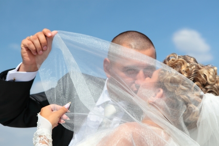 The groom and the bride passionately kiss under a veil against the blue sky in a sunny dayの写真素材