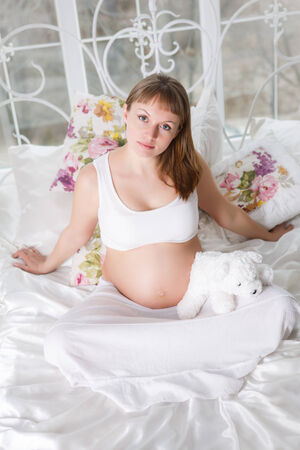 Beautiful pregnant woman posing while siting on a bed at homeの写真素材