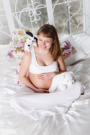 Beautiful pregnant woman posing while siting on a bed at homeの写真素材