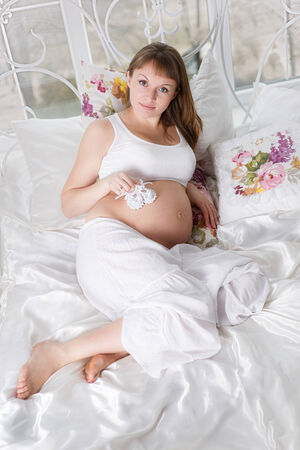Beautiful pregnant woman with children s bootees posing while lying on a bed at homeの写真素材