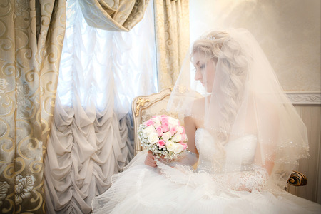 Portrait of the bride with a wedding bouquet from roses in the hand sitting on a sofa near a windowの写真素材