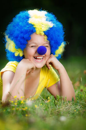 Smiling girl in clown wig with blue nose is lying on the green grass in the parkの写真素材