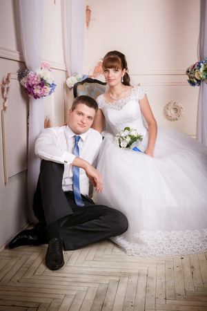 The bride sits in a chair and the groom sits near bride on the floor in the room with a beautiful interiorの写真素材