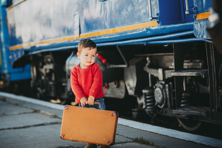 Adorable little kid boy dressed in red sweater on a railway station near train with retro old  brown suitcase. Ready for vacation. Young traveler on the platform.の写真素材