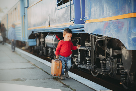 Adorable little kid boy dressed in red sweater on a railway station near train with retro old  brown suitcase. Ready for vacation. Young traveler on the platform.の写真素材