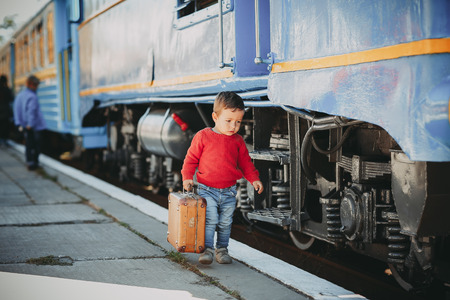 Adorable little kid boy dressed in red sweater on a railway station near train with retro old  brown suitcase. Ready for vacation. Young traveler on the platform.の写真素材