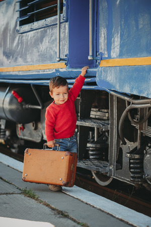 Adorable little kid boy dressed in red sweater on a railway station near train with retro old  brown suitcase. Ready for vacation. Young traveler on the platform.の写真素材
