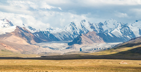 Yellow fields stretch out in front of rocky mountains,tops of which are covered with snow.The ridges rise in the distance, creating a contrast between the warm shades of the fields and the cold peaksの写真素材