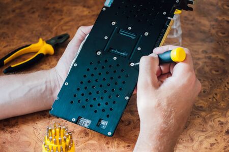 Computer video card repair. Male hands with a yellow screwdriver on a wooden backgroundの写真素材