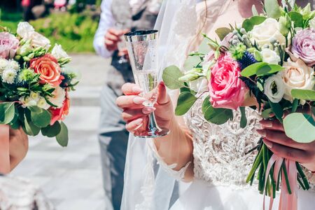 A bride with long nude nails is holding a glass of champagne. Against the background of the bridal bouquet and bridesmaid bouquet. Close-upの写真素材