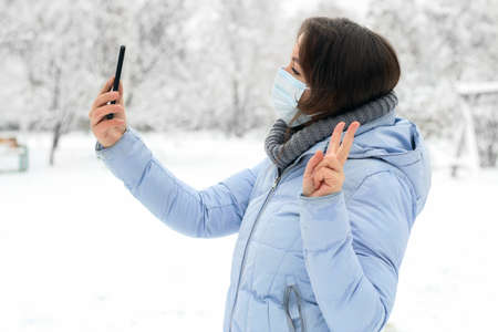A girl in a medical mask in the winter outdoors on the background of snow makes a selfie on the phone.の写真素材