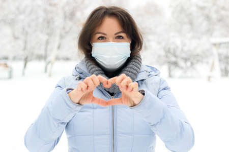 A girl in a medical mask in the winter outdoors on a background of snow makes a heart with her handsの写真素材