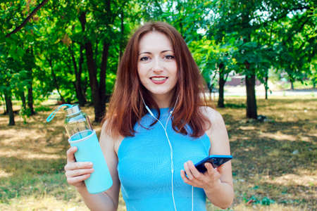 A young pretty girl with red hair looks at the camera, put on headphones, holding water in her hands. A girl listening to a podcast about a healthy lifestyle.の写真素材