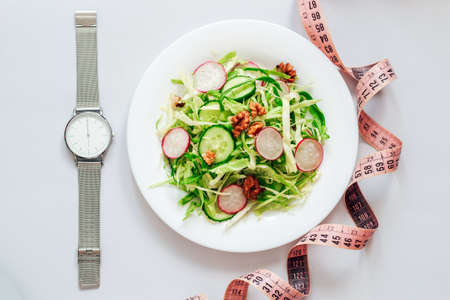 Cucumber salad with radishes, cabbage and nuts on a white plate. Hours with a time of 6 pm so as not to eat. Top view on gray background.の写真素材