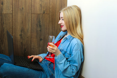 Girl with blond hair in denim clothes sits with a glass of champagne with a laptop.の写真素材