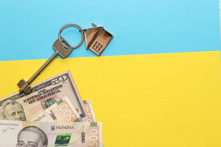 The key to the apartment with a keychain in the shape of a house on a cardboard blue yellow background, the colors of the flag of Ukraine, hryvnias and dollars.の写真素材