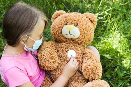 A girl in a medical mask plays doctor with a toy bear.の写真素材