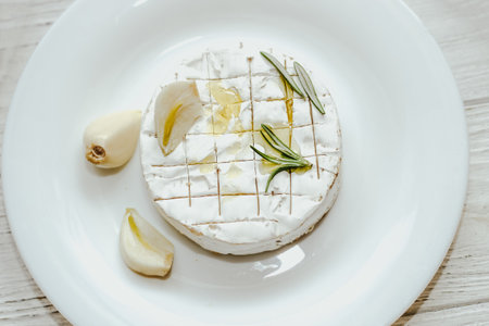 Round cheese in camembert mold with a sprig of rosemary, with garlic on the table.の写真素材