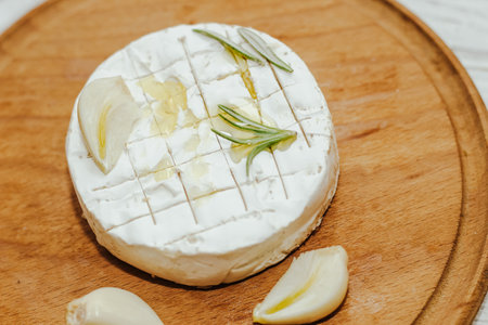 Round cheese in camembert mold with a sprig of rosemary, with garlic on the table.の写真素材