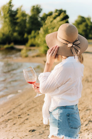 A girl in a straw hat with a glass of wine sits on the beach at sunset.の写真素材