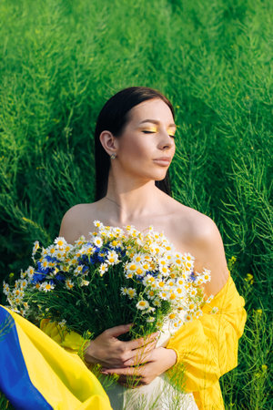 Ukrainian girl with a blue-yellow flag in her hands and a bouquet of flowers on the background of the fieldの写真素材