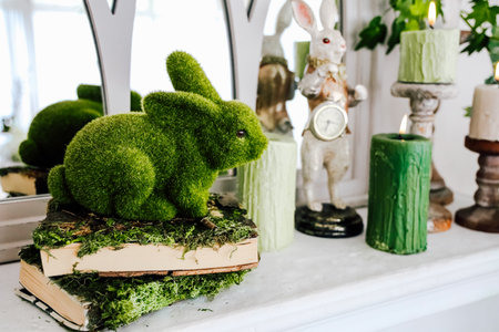 A green moss-covered bunny sits on books, surrounded by decorative candles and an elegant mirror in a tranquil room.の写真素材