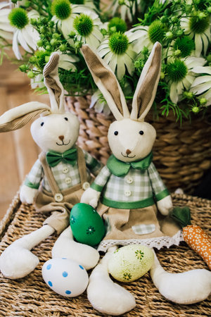 Two charming plush bunnies sit with brightly colored Easter eggs next to a basket of flowers, creating a festive atmosphere.の写真素材