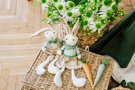 Soft fabric bunnies dressed in spring attire sit on a woven basket surrounded by vibrant flowers and carrots.の写真素材