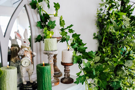 Candles in various colors are arranged on a shelf surrounded by lush greenery and a rabbit figurine,...