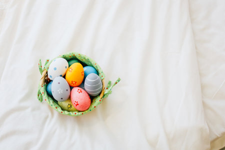 Brightly colored eggs are neatly placed in a green basket resting on a white blanket, creating a cheerful spring setting.の写真素材