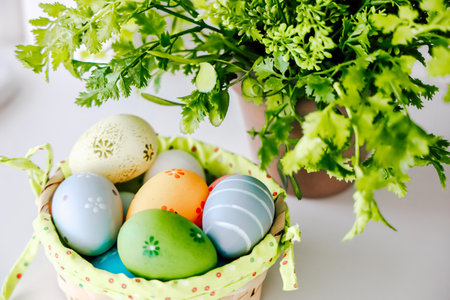 Vibrant Easter eggs in pastel colors rest in a woven basket next to a pot of fresh herbs on a sunlit table.の写真素材