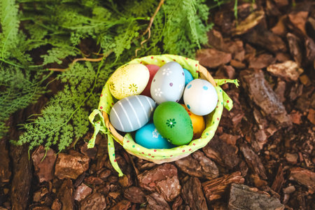 A basket filled with vibrant, decorated Easter eggs is placed in a natural setting with green...