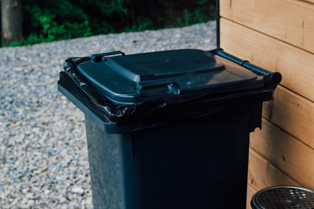 A dark trash bin stands beside a wooden wall surrounded by gravel reflecting a calm moment in a natural setting.の写真素材