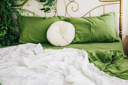 A comfortable bed showcases lush green sheets and a round decorative pillow, framed by soft white bedding and indoor plants nearby.の写真素材